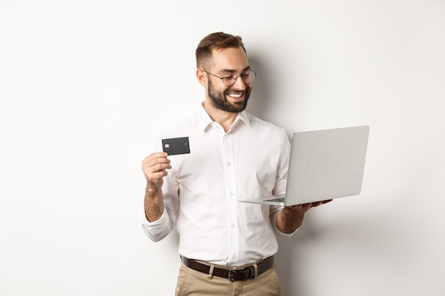 Compras online. Homem de negócios bonito segurando um cartão de crédito e usando laptop, fazendo pagamento pela internet, em pé sobre um fundo branco