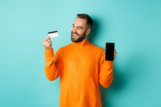 Compras online. Homem satisfeito usando cartão de crédito e mostrando a tela do celular, parecendo satisfeito