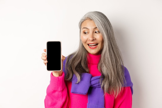 Compras online. Linda e elegante mulher asiática sênior com cabelo grisalho, mostrando a tela do smartphone em branco e parecendo satisfeita, em pé sobre um fundo branco