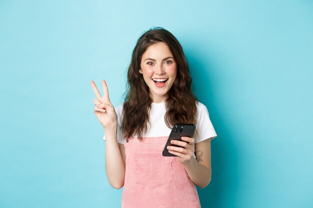 Compras online. menina sorridente positiva mostrando o sinal-v e olhando feliz para a câmera, usando o smartphone, segurando o telefone móvel, em pé contra um fundo azul.