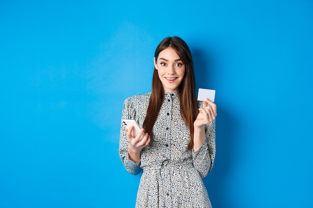 Compras online. Mulher bonita, pagando o pedido com smartphone e cartão de crédito de plástico, sorrindo para a câmera, usando vestido azul.