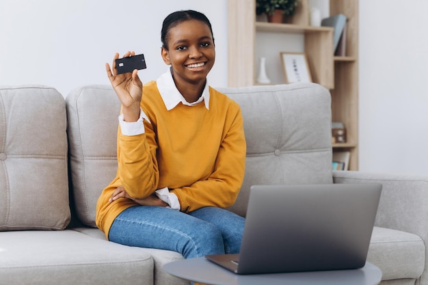 Compras online. Positiva jovem negra usando laptop e cartão de crédito em casa, fazendo compras na Internet, sentada no sofá na sala de estar