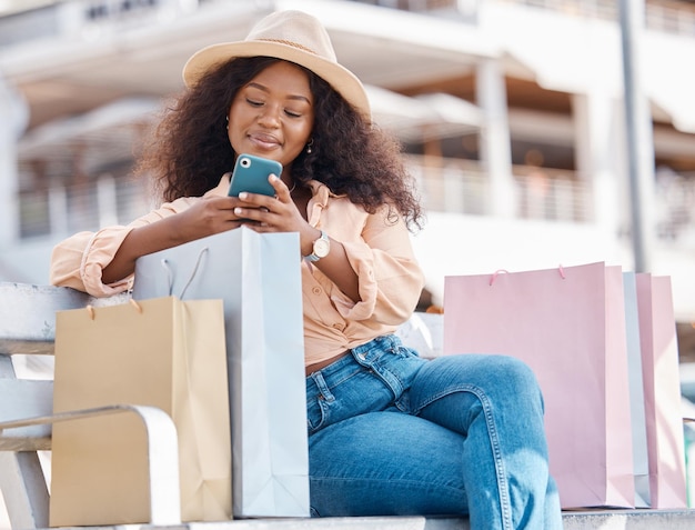 Compras por telefone e mulher negra feliz com sacolas de compras e lojas em um banco ao ar livre Rolagem de aplicativo de comércio eletrônico e móvel on-line de um sorriso de jovem de Miami com tecnologia e sacola de papel de varejo