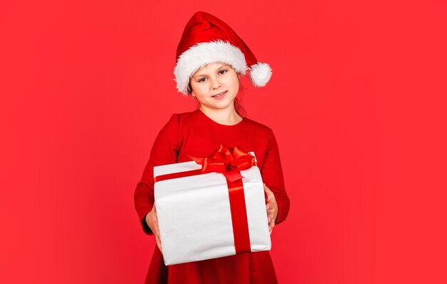 Compras de regalos de Navidad. Paquete de regalo. Fines promocionales. Donar. Feliz Navidad y felices fiestas. Vacaciones de invierno. Caja de regalo del asimiento del sombrero de Santa de la niña. Niño mantenga presente cuadro de fondo rojo.