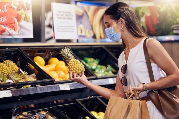 Compras en el supermercado revisando y sosteniendo piña con una mujer mirando saludable para comer Mujer joven con una máscara covid comprando fruta fresca y comida en una tienda de comestibles durante una pandemia