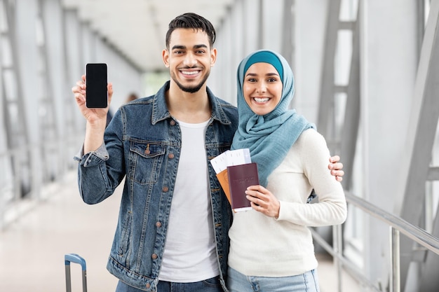 Compre bilhetes online casal muçulmano feliz mostrando smartphone em branco no aeroporto