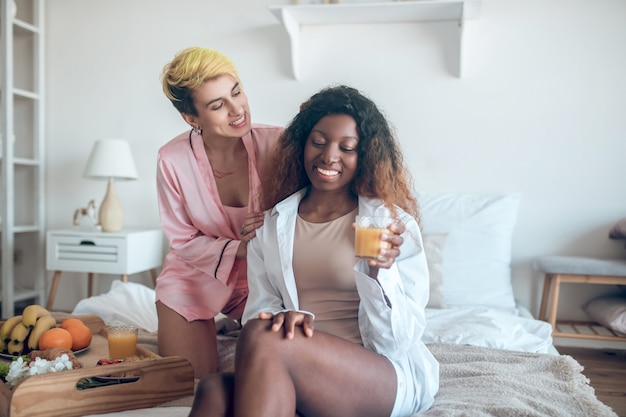 Comprensión. Dos mujeres bonitas jóvenes brillantes en ropa de dormir charlando felices en la cama durante el desayuno ligero