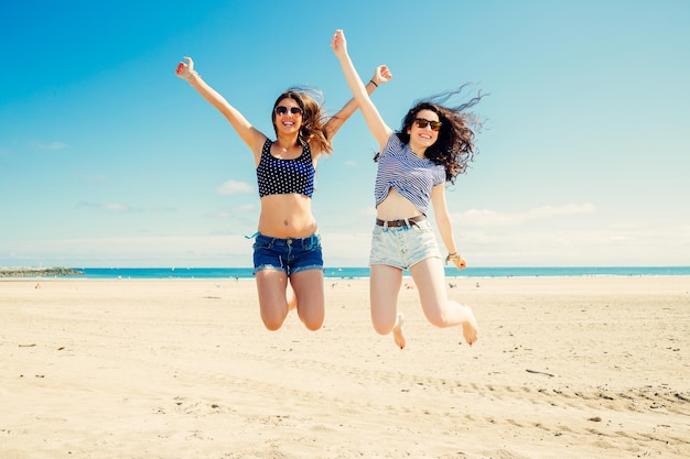 Comprimento completo de amigas excitadas saltando na praia durante o dia ensolarado