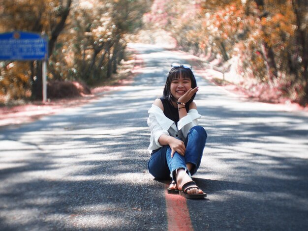 Foto comprimento completo de jovem alegre sentada na estrada na floresta durante o outono