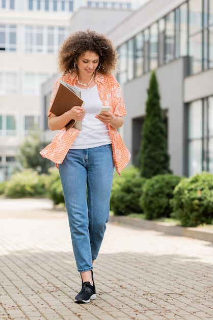 comprimento completo de mulher alegre com cabelo encaracolado usando smartphone e segurando laptop com pasta enquanto