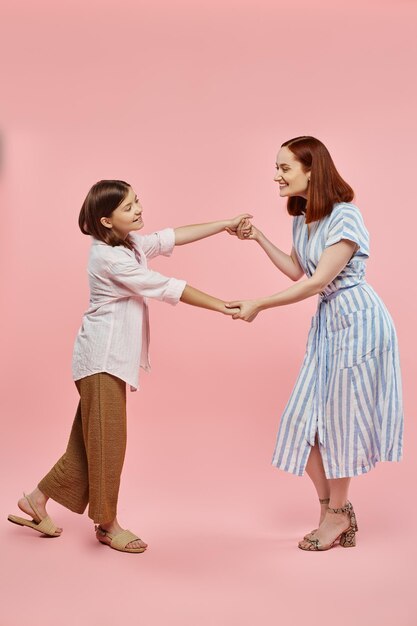 comprimento completo de mulher despreocupada e elegante com filha pré-adolescente de mãos dadas e dançando em rosa