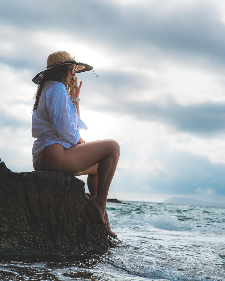 Foto comprimento completo de mulher usando chapéu sentada em uma rocha contra o mar e o céu