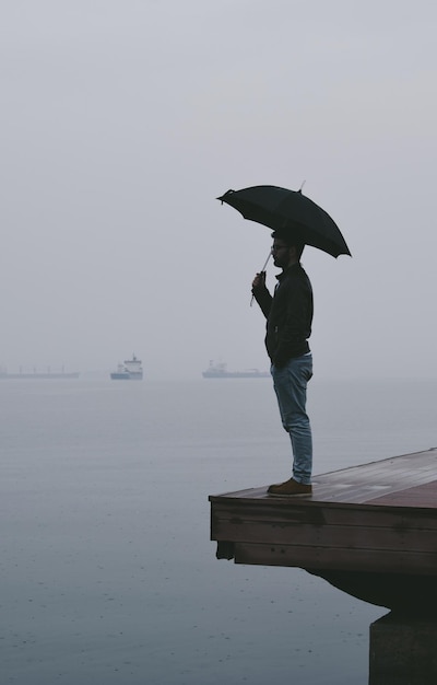 Comprimento completo de um jovem segurando um guarda-chuva enquanto está de pé no cais sobre o mar contra o céu