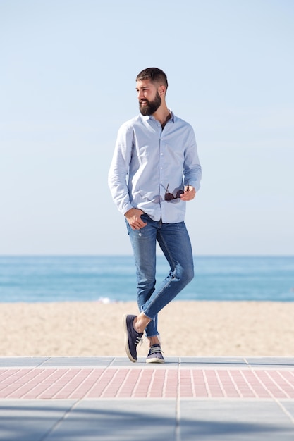 Comprimento total bonito homem segurando óculos de sol e em pé na praia