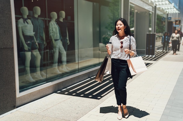 comprimento total da bela jovem asiática carregando sacola de compras. Linda garota andando em uma rua da cidade depois de fazer compras na venda de verão no shopping center em stanford. senhora segurando a xícara de café perto da janela.