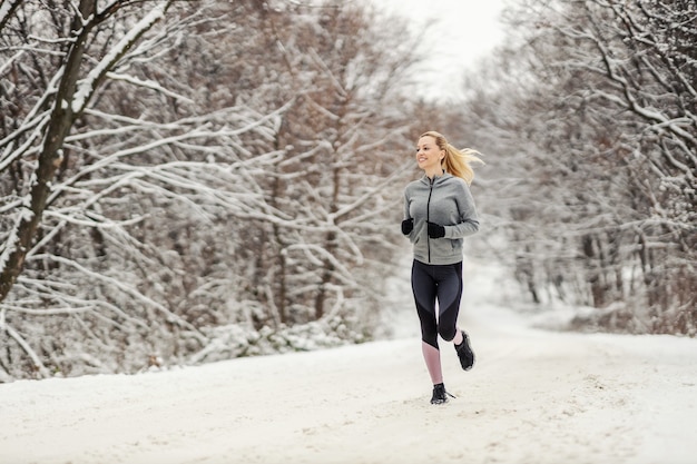 Comprimento total da mulher de meia-idade feliz correndo na natureza no caminho de neve no inverno. Aptidão de inverno, hábitos saudáveis