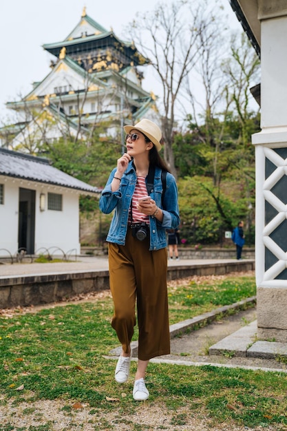 comprimento total da senhora viajante com chapéu de óculos de sol visita o castelo de osaka. jovem turista segurando celular pesquisando no aplicativo de mapa online andando no jardim. viajar sozinho no conceito de férias de verão.