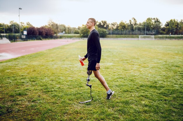 Comprimento total de bonito apto homem deficiente caucasiano desportivo em roupas esportivas e com perna artificial andando na quadra de futebol e segurando refrescos.