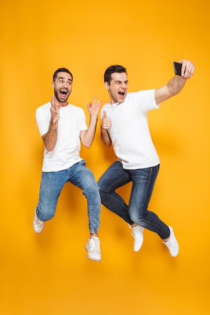 Comprimento total de dois amigos homens alegres e animados, vestindo camisetas em branco, pulando isolados sobre a parede amarela, tirando uma selfie