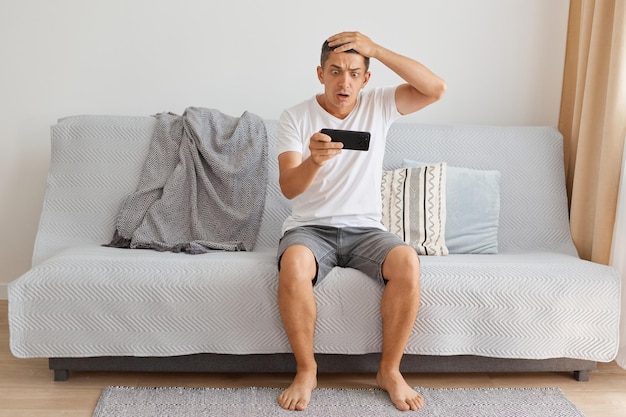 Comprimento total de homem de cabelo escuro caucasiano chocado, vestindo camiseta branca estilo casual, sentado na tosse jogando videogame, ficando surpreso com o resultado do nível, mantendo a mão na cabeça