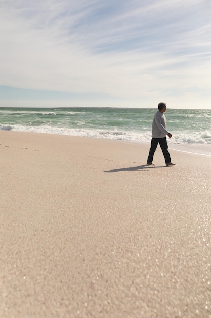 Comprimento total de homem sênior birracial aposentado andando na praia contra o céu durante o dia ensolarado