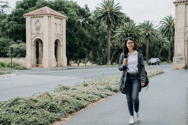 comprimento total de jovem estudante de intercâmbio vai ter aula andando na universidade. garota de faculdade asiática segurando café e bolsa de transporte em stanford. senhora inteligente trabalhando duro na escola indo para o curso.