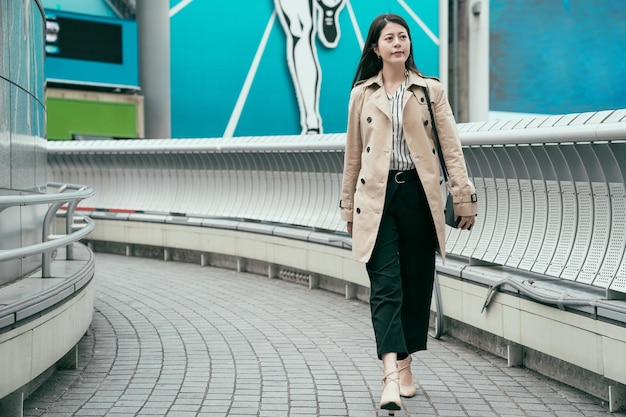 comprimento total de trabalhador menina asiática confiante andando na ponte ao pôr do sol indo para casa do trabalho. jovem mulher japonesa cruzando viaduto usando sapatos de salto alto. mulher sorridente carregando bolsa pequena.