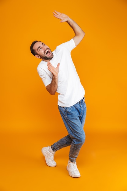 Comprimento total de um cara alegre em camiseta e jeans levantando as mãos em pé, isolado no amarelo