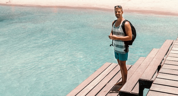 Comprimento total de um homem bonito branco com mochila e roupas de verão, usando telefone inteligente em pé no cais. Conceito de férias de verão.