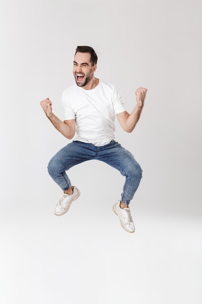 Comprimento total de um homem bonito e alegre vestindo uma camiseta em branco, pulando isolado na parede branca