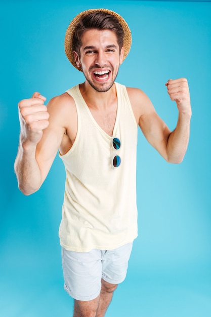 Comprimento total de um jovem alegre de chapéu e shorts em pé e comemorando o sucesso na parede azul