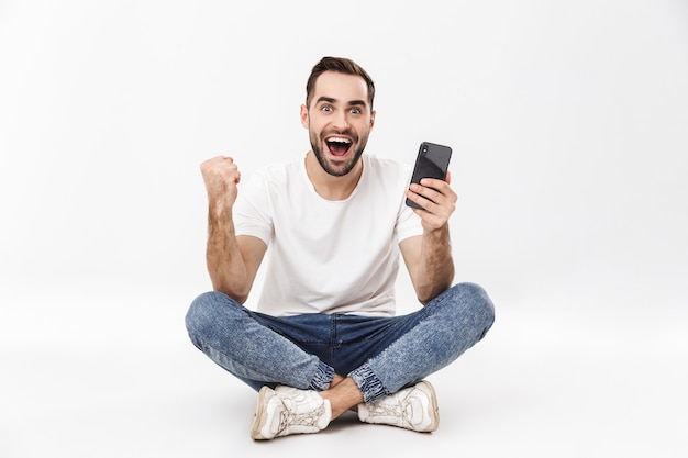 Comprimento total de um jovem alegre sentado com as pernas cruzadas isoladas sobre uma parede branca, usando telefone celular, comemorando