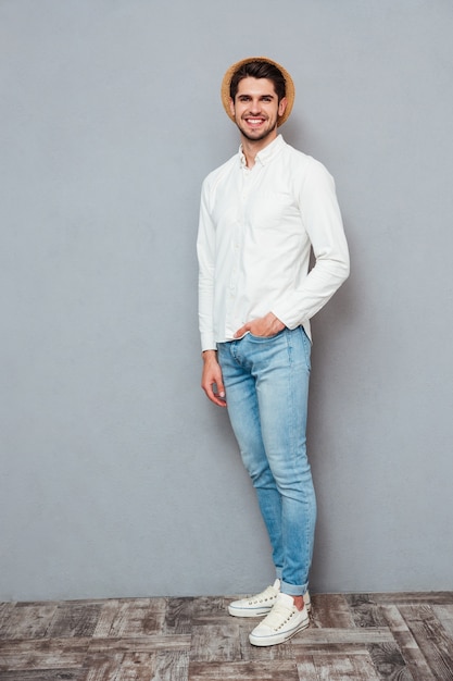 Comprimento total de um jovem bonito feliz de camisa branca, jeans e chapéu em pé