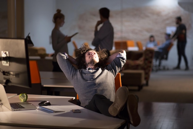 Foto comprimento total de um jovem empresário casual relaxado sentado com as pernas na mesa no escritório noturno
