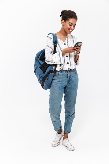 Comprimento total de um retrato de uma jovem africana atraente carregando uma mochila em pé, isolada na parede branca, usando telefone celular
