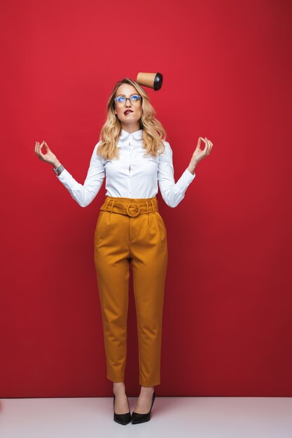 Foto comprimento total de uma bela jovem loira em pé isolado sobre um fundo vermelho, equilibrando-se, segurando a xícara de café