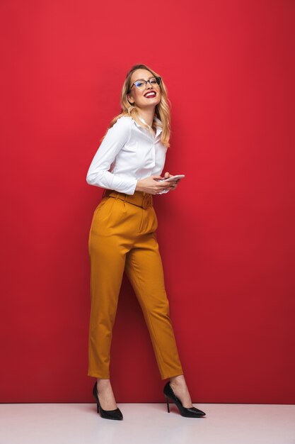 Comprimento total de uma bela jovem loira feliz em pé isolado sobre um fundo vermelho, segurando um telefone celular