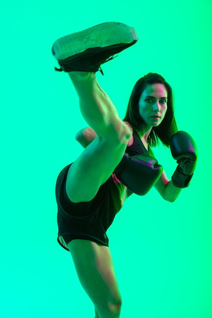 Comprimento total de uma linda garota jovem e confiante em fitness, em pé, isolada sobre uma parede de néon verde, chutando com a perna durante o boxe