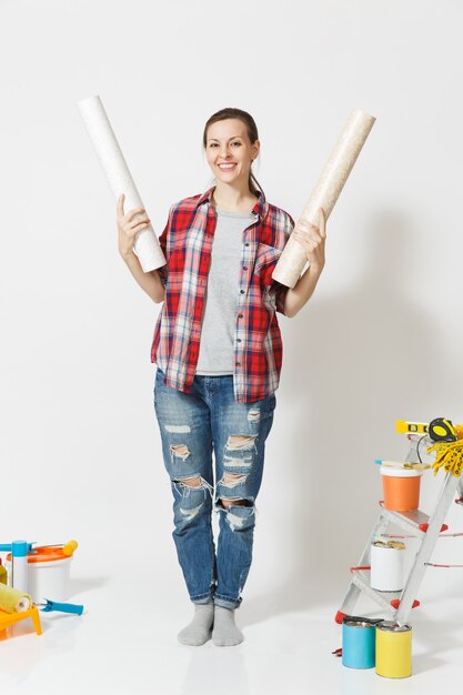 Comprimento total de uma linda mulher com roupas casuais em pé perto de instrumentos para casa de apartamento de renovação isolado no fundo branco. Papel de parede, acessórios para colar, ferramentas de pintura. Conceito de reparo.