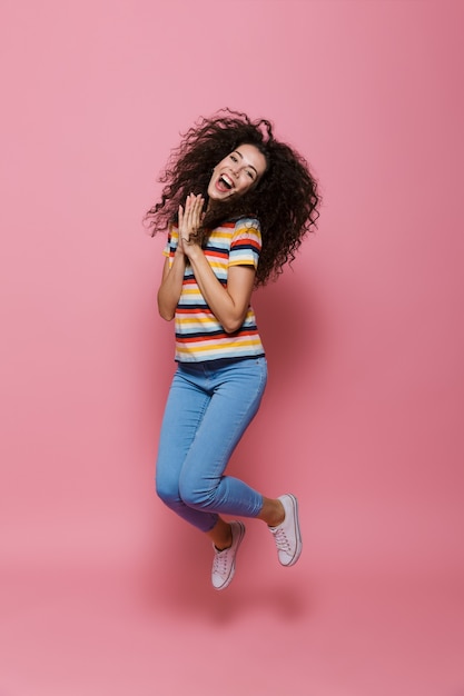 Comprimento total de uma linda mulher de 20 anos com cabelo encaracolado se divertindo e pulando isolado na rosa