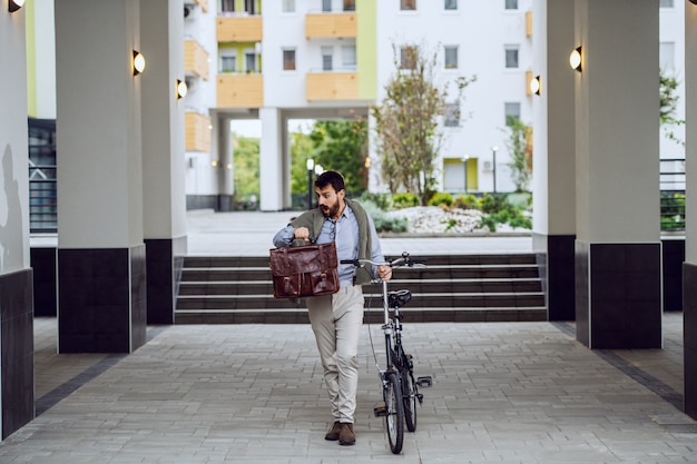 Comprimento total do empresário caucasiano elegante bonito segurando sua mala, empurrando a bicicleta, olhando para o relógio de pulso e indo para o trabalho.