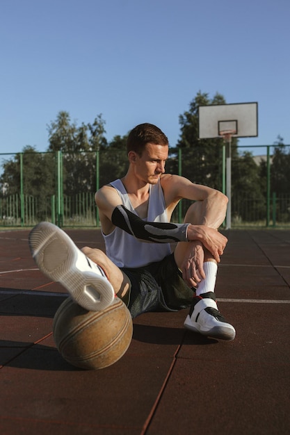 Foto comprimento total do jogador masculino em forma com basquete sentado no playground no verão e olhando para longe