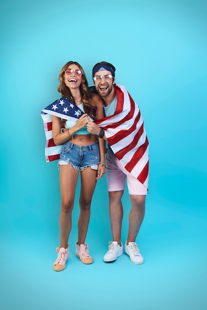 Comprimento total do lindo jovem casal coberto com a bandeira americana, sorrindo em pé contra o azul.