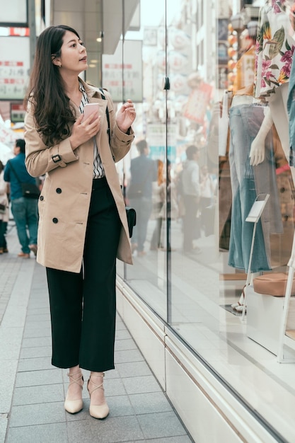 comprimento total jovem mulher asiática com celular olhando para a vitrine andando na área comercial em dotonbori osaka japão. relaxe linda viciada em compras aproveite o tempo livre com telefone inteligente na rua.