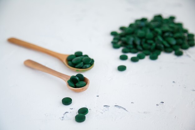 Comprimidos de espirulina verdes sobre um fundo branco de concreto. vários comprimidos em colheres de madeira