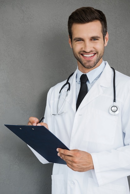 Foto comprobando el resultado. feliz joven médico en uniforme blanco mirando a cámara y sosteniendo el portapapeles mientras está de pie contra el fondo gris