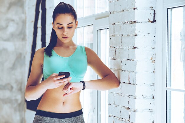 Foto comprobando todas las actualizaciones de formación. hermosa joven mirando su pulsera deportiva y usando su teléfono inteligente mientras se inclina a la pared en el gimnasio