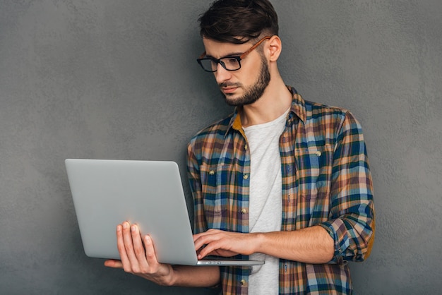 Comprobando todas las actualizaciones. Hombre joven confidente usando su computadora portátil y mirando concentrado mientras está de pie contra el fondo gris