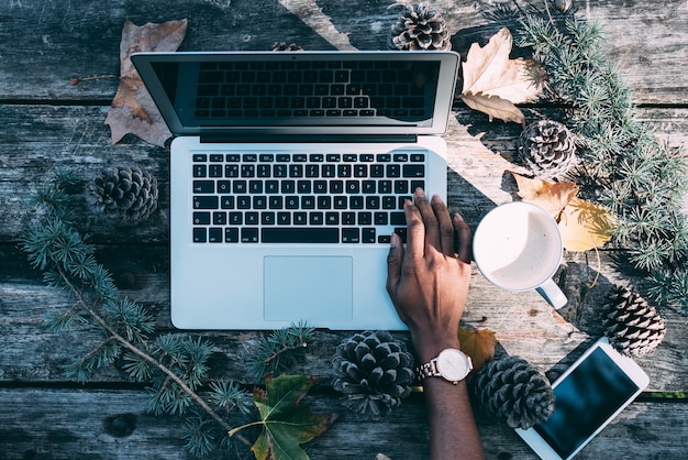 Computador em uma mesa de madeira com café e pinheiros ao ar livre