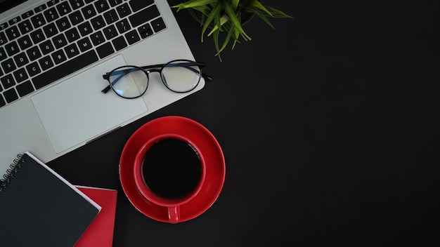 Computador laptop, xícara de café vermelha, óculos e caderno na mesa preta